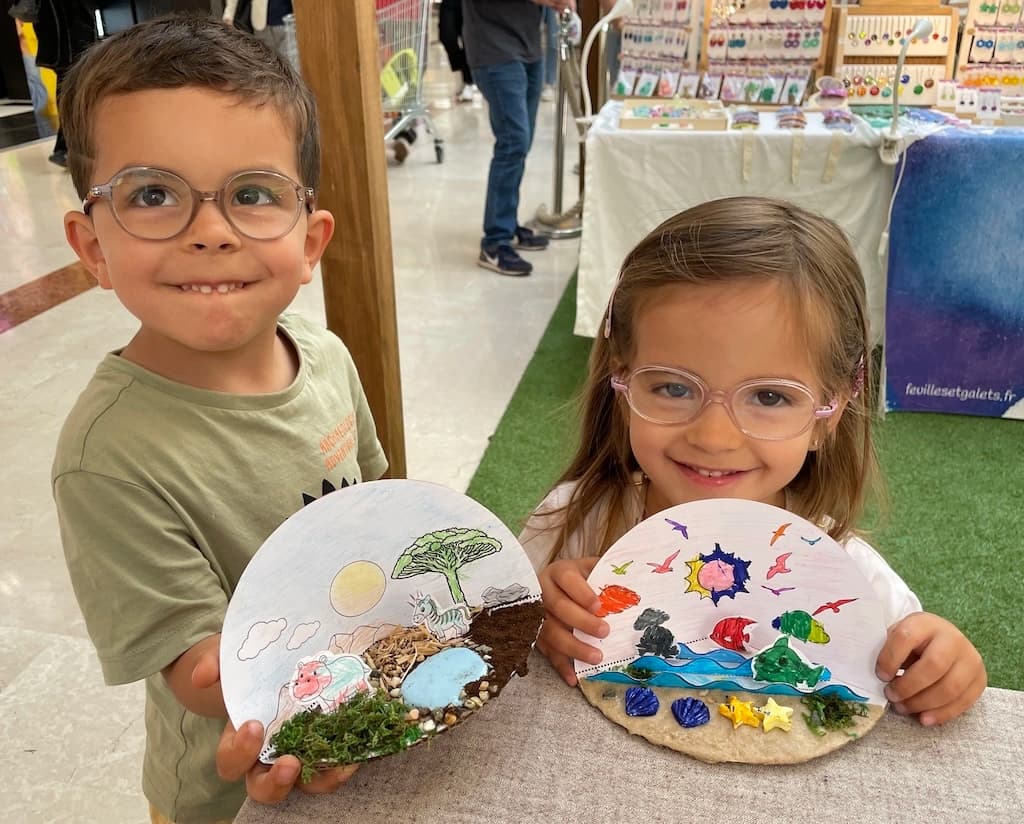 Deux enfants souriants montrent leurs créations : un paysage de savane et un fond marin sur des disques en carton