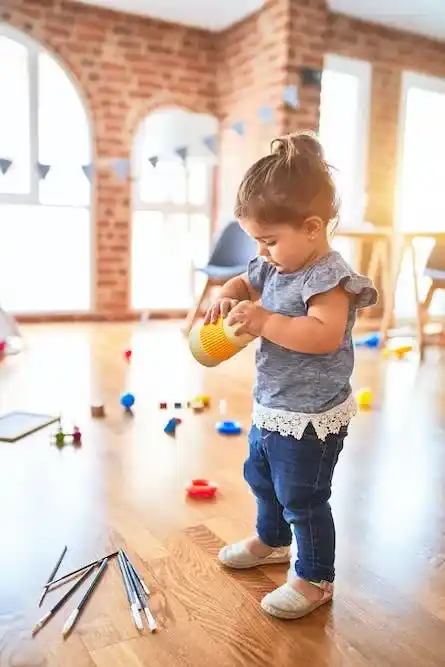 Une petite fille joue avec un gobelet dans une salle lumineuse, entourée de crayons et de jouets